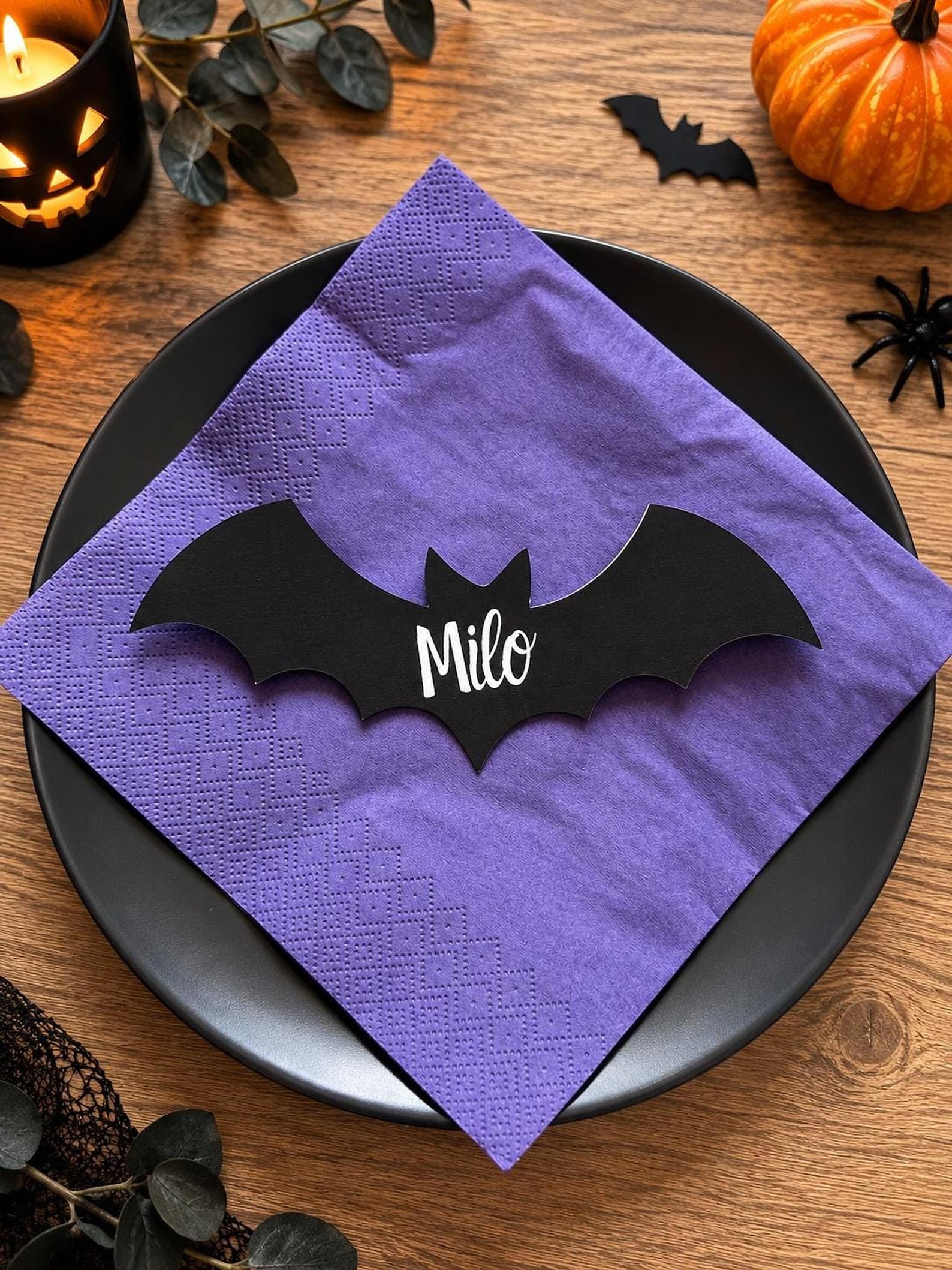 A Black Bat Laid Across the Napkin Black bat-shaped place card with a white name across its center.
