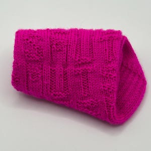 Könnte beinhalten: Ein leuchtend pinkfarbenes Strickteil, möglicherweise ein Schal oder ein Kragen, mit einem strukturierten Muster. Das Strickwerk ist gefaltet und zeigt das komplizierte Stichdesign und die satte, gesättigte Farbe. Der Artikel wirkt weich und warm.
