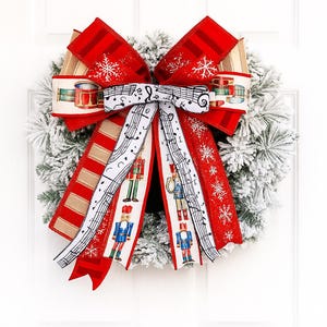 May include: A festive Christmas wreath with a large red bow. The bow features ribbons with snowflake, musical note, and nutcracker soldier designs. The wreath is covered in faux snow, creating a wintery holiday decoration.