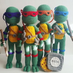 May include: Four crocheted Teenage Mutant Ninja Turtles, each with a different colored mask and weapon. The turtles are standing in a row, with a pizza box and a slice of pizza in front of them.