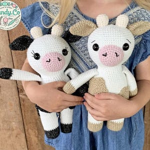 Op de afbeelding: Twee gehaakte knuffels, een zwart-wit en een bruin-wit, worden vastgehouden door een kind. De koeien hebben zwarte ogen, roze neuzen en wit garen voor hun lichamen.