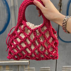 May include: A bright pink crocheted mesh bag with a beige fabric lining. The bag has a single top handle and a drawstring closure.