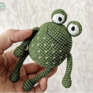 Op de afbeelding: Een groene gehaakte kikker met witte ogen en een textuur. De kikker heeft lange armen en benen.