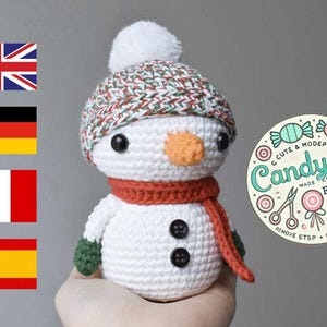 Puede incluir: Un juguete amigurumi de muñeco de nieve tejido a crochet con un gorro a rayas rojo y verde, una bufanda naranja y ojos de botón negros. El muñeco de nieve sostiene una manopla de crochet verde. El fondo incluye un logotipo para la tienda Etsy Candy.Co.