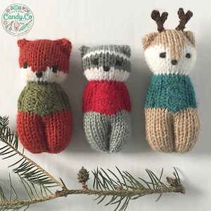 Può includere: Tre giocattoli di animali a maglia: una volpe con un maglione verde, un procione con un maglione rosso e una renna con un maglione turchese. I giocattoli sono seduti su una superficie bianca con un ramo di pino in primo piano.