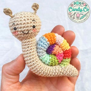 Könnte beinhalten: Ein gehäkeltes Amigurumi-Spielzeug in Form einer Schnecke mit einem beigefarbenen Körper und einer bunten Regenbogenmuschel. Die Schnecke hat ein lächelndes Gesicht mit schwarzen Augen und rosa Wangen.
