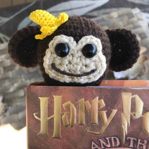 Puede incluir: Un marcapáginas de ganchillo con forma de mono con un plátano amarillo en la cabeza. El marcapáginas está sobre un libro titulado "Harry Potter y..."