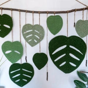 Op de afbeelding: Een wanddecoratie gemaakt van gecrochete groene bladeren in de vorm van harten. De bladeren hangen aan een tak met touw en kralen.