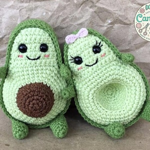 Puede incluir: Dos peluches de aguacate de crochet con caras sonrientes. Un aguacate tiene un lazo rosa en la cabeza. Los aguacates son verdes con hoyos marrones.