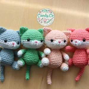 Może przedstawiać: Cztery pluszowe zabawki w kształcie kota wykonane na szydełku w różnych kolorach: jasnoniebieskim, zielonym, różowym i koralowym. Każdy kot ma białe cechy twarzy i czarne oczy.