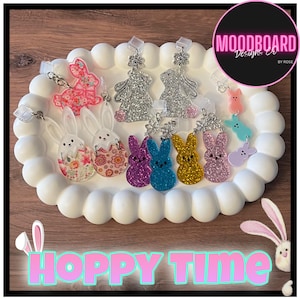 May include: A collection of Easter-themed earrings and charms in various colors and designs, including glittery bunnies and floral egg designs. The earrings are displayed on a white, decorative tray. The image also includes the text "HOPPY TIME".