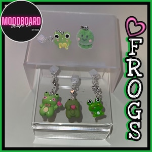 May include: A display of frog-themed earrings. The earrings are in various shades of green, with some featuring heart and flower charms. The word "FROGS" is displayed vertically in a playful font. The brand logo "Moodboard Designs Co" is also visible.