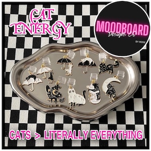May include: A collection of cat-themed earrings displayed on a silver tray. The earrings feature black and white cat designs, including cats under umbrellas, hugging, and on moons. The image includes the text "CAT ENERGY" and "CATS > LITERALLY EVERYTHING."