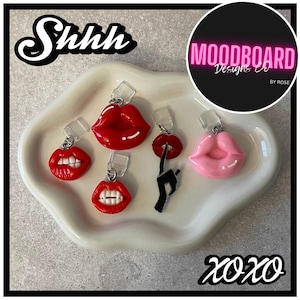 May include: A collection of lip-shaped charms in red and pink, displayed on a white, wavy-edged dish. The charms vary in design, some with open mouths and visible teeth. The image includes the text "Shhh" and "xoxo".