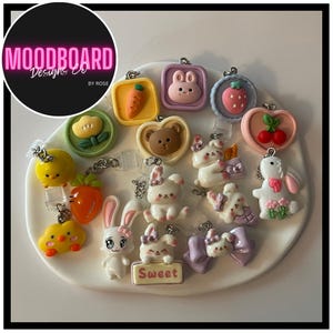 May include: A collection of colorful, decorative charms featuring various designs, including a carrot, bear, bunny, chick, strawberry, and bow. The charms are attached to keychains and are arranged on a white surface.