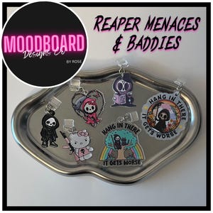 May include: A collection of acrylic charms featuring grim reaper designs and the text "Hang in there, it gets worse." Charms include a grim reaper reading a book, a reaper in a heart, and a reaper with a weapon. The charms are on a silver tray.