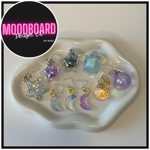 May include: A collection of iridescent charms in various shapes, including stars, moons, and spheres, displayed in a white, cloud-shaped dish. The charms have silver and gold accents. The image includes a logo that says "MOODBOARD Designs Co BY ROSE".