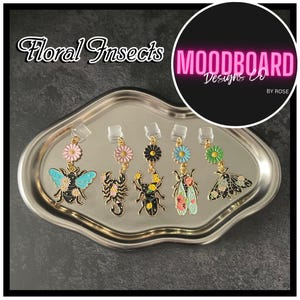 May include: A collection of floral insect earrings displayed on a silver tray. Each earring features a different insect design, including a bee, scorpion, beetle, cicada, and moth, adorned with colorful floral accents. The text "Floral Insects" is visible.