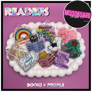 May include: A collection of clear acrylic charms featuring various book-themed illustrations and text, including phrases like 'Just one more chapter', 'A book a day keeps reality away', and 'Books are better than people', arranged on a white scalloped tray.