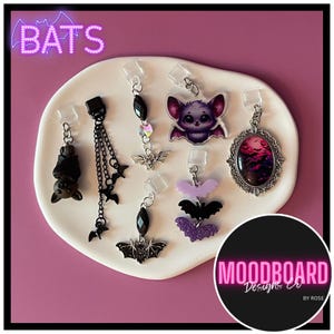 May include: A collection of six bat-themed charms featuring various designs including a hanging black bat, a multi-chain bat dangle, a crystal-accented bat, a purple bat character, a layered bat stack, and an oval pendant with a bat-filled sky scene.