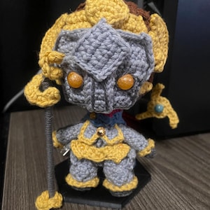 May include: A crocheted figurine of a warrior wearing a golden crown and armour, holding a staff with a yellow orb at the top. The figurine has glowing orange eyes and is standing on a black base.