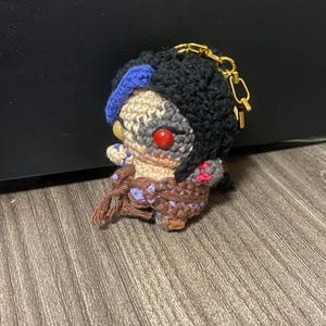 May include: Crocheted keychain featuring a character with black hair, a blue headband, and red eyes. The character is wearing a brown and purple outfit and has a gold chain attached.