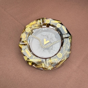 Puede incluir: Un cenicero redondo con un exterior de resina transparente incrustado con casquillos de bala dorados. El centro presenta un diseño gris oscuro con una cadena dorada y la letra "L" dorada.