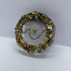Puede incluir: Un cenicero octogonal transparente con casquillos de bala de latón incrustados en resina. Una cadena dorada y un emblema "V" dorado están presentes. El cenicero está diseñado para el uso de tabaco y fines decorativos.