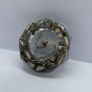 Puede incluir: Un cenicero de resina transparente con un centro circular y una forma exterior octogonal. El cenicero está lleno de casquillos de bala plateados. Una cadena plateada y la letra "V" están en el centro.