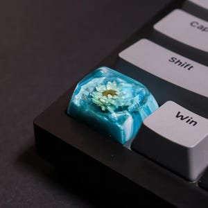 Peut inclure: Une touche de clavier personnalisée avec une petite fleur vert clair encapsulée dans de la résine bleue translucide. La touche est placée sur un clavier noir, avec les touches "Shift" et "Win" visibles.