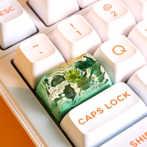 May include: A green and white resin keycap with a swirling pattern, sitting on a white keyboard with the "Caps Lock" key visible.