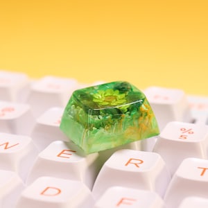 Peut inclure: Une touche de clavier personnalisée avec un motif floral vert et jaune encapsulé dans de la résine transparente. La touche est posée sur un clavier blanc avec un fond jaune. La touche a un aspect artistique unique.