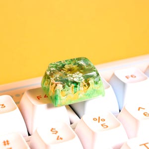 Peut inclure: Un capuchon de touche en résine verte et dorée avec une fleur pressée à l'intérieur. Le capuchon de touche est posé sur un clavier blanc avec des lettres orange.