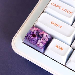 Peut inclure: Une touche de clavier personnalisée avec un motif floral. La touche est de couleur violet clair avec des fleurs violettes et roses incrustées. Les touches environnantes sont blanches avec des lettres orange, dont "CAPS LOCK", "SHIFT" et "WIN".