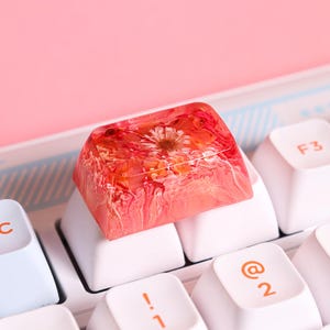 May include: A pink and white resin keycap with a pressed flower design. The keycap is sitting on a white keyboard.