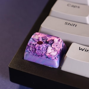 Peut inclure: Une touche de clavier personnalisée avec un motif floral. La touche présente un mélange de couleurs violet, rose et blanc, avec une fleur violet foncé au centre. La touche est placée sur un cadre de clavier noir et des touches grises.