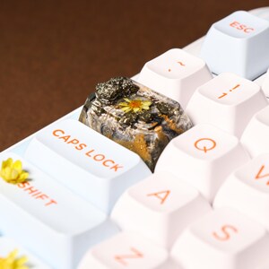 May include: Close-up of a custom keyboard with white keys and a unique artisan keycap. The keycap is a clear resin with a rock-like appearance, featuring a yellow flower and mineral inclusions. The keyboard keys are labeled with orange text.