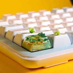 Peut inclure: Gros plan sur un clavier mécanique avec des touches blanches et une touche artisanale personnalisée. La touche artisanale est en résine translucide verte et jaune avec un motif de paysage. De petites plantes vertes sont placées sur certaines touches.