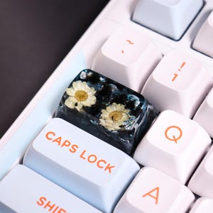 May include: Close-up of a custom keyboard with a unique resin keycap. The keycap features two small white and yellow flowers encased in a dark blue and black resin. The keyboard has white keys with orange lettering.