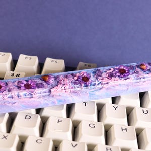 Könnte beinhalten: Eine individuelle Leertaste für die Tastatur mit floralem Design. Die Tastenkappe ist transparent mit einem lila und blauen Marmoreffekt und verfügt über eingebettete lila Blumen mit orangefarbenen Zentren. Die Tastenkappe liegt auf einer weißen Tastatur.