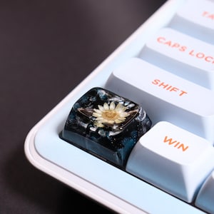 Peut inclure: Gros plan sur une touche de clavier personnalisée avec une fleur séchée encapsulée dans de la résine. La touche est noire et bleue avec une fleur blanche et jaune. Les touches du clavier sont bleu clair et blanches.