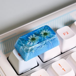May include: A blue resin keycap with two white flowers pressed inside. The keycap is on a white keyboard with a blue and white striped background.