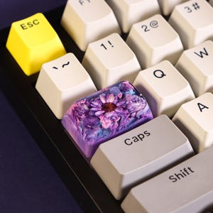 May include: Close-up of a mechanical keyboard with a custom keycap. The keycap features a purple and blue resin design with a flower. Other keys are white with black lettering, including "ESC", "Caps", and "Shift". The ESC key is yellow.