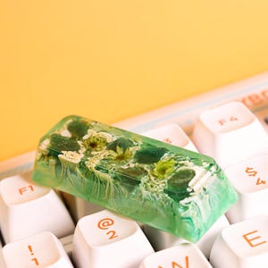 Könnte beinhalten: Eine grüne Harztastenkappe mit eingebetteten getrockneten Blumen. Die Tastenkappe liegt auf einer weißen Tastatur mit Tasten, die mit "F1", "@2" und "E" beschriftet sind.
