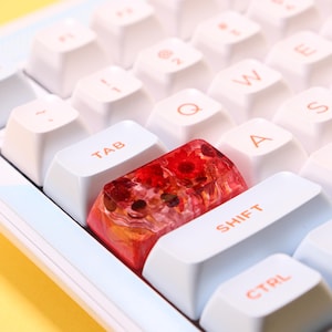 May include: Close-up of a white keyboard with a custom keycap. The keycap is transparent with red flowers and other botanical elements encased within. The other keys are white with orange lettering.