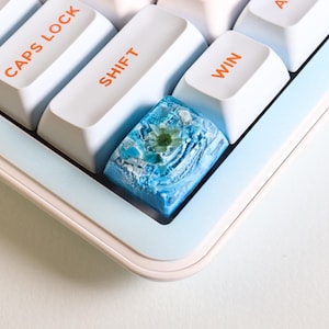 Peut inclure: Gros plan sur un clavier personnalisé avec des touches blanches et bleu clair. Les touches "SHIFT", "CAPS LOCK" et "WIN" sont visibles. Une touche en résine bleue unique avec un motif floral est présentée, ajoutant une touche décorative au clavier.