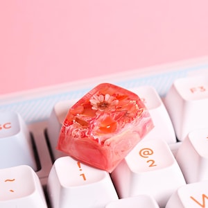 Peut inclure: Un capuchon de touche en résine rose avec une fleur séchée intégrée. Le capuchon de touche est posé sur un clavier blanc avec des touches étiquetées "!", "@" et "2".
