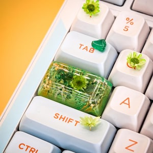 May include: A white keyboard with green and gold resin keycaps featuring pressed flowers. The keycaps are labeled "SHIFT", "TAB", "A", and "Z".