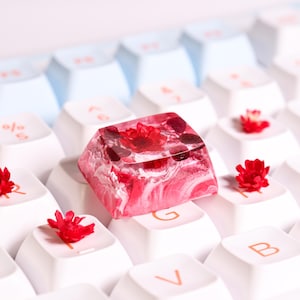 Peut inclure: Un capuchon de touche en résine rose et blanche avec un motif de fleurs pressées. Le capuchon de touche est posé sur un clavier blanc avec d'autres touches qui ont des fleurs pressées.