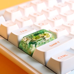 May include: A white keyboard with a green resin keycap featuring a floral design. The keycap is labeled "SHIFT".